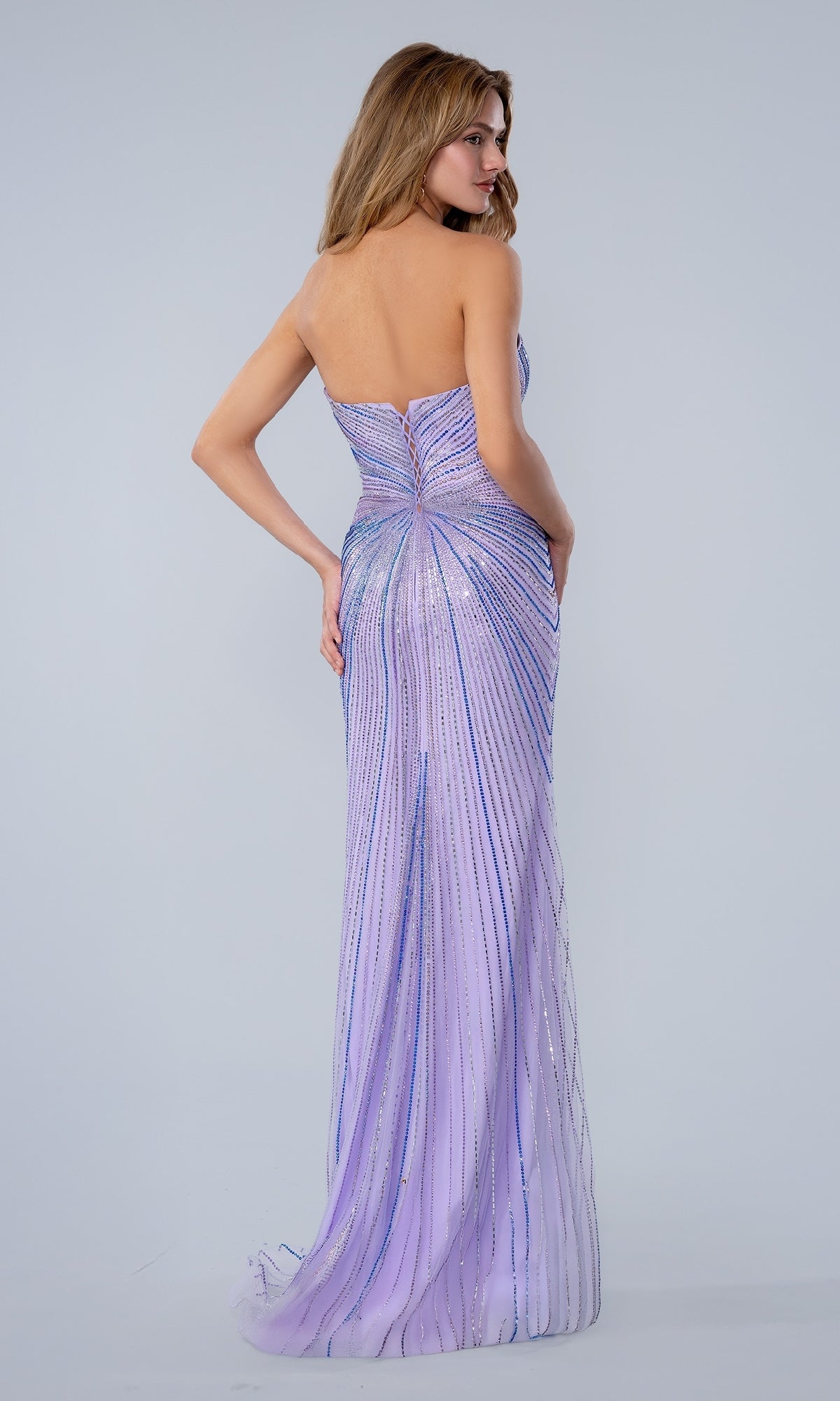 A woman faces away, wearing the Stella Couture 26163 Long Prom Dress—a strapless lavender gown with a fitted silhouette, shimmering vertical stripes, and a floor-length hem.